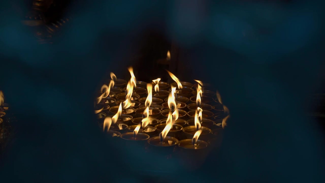 Close up static shot of numerous burning butter lamps with bright flames flickering in a dark temple room, viewed through an artistic blurred out-of-focus background frame in Kathmandu, Nepal.