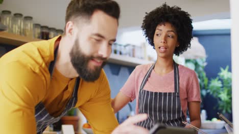 Video-of-happy-diverse-couple-in-aprons-looking-at-tablet-before-preparing-food-in-kitchen-at-home