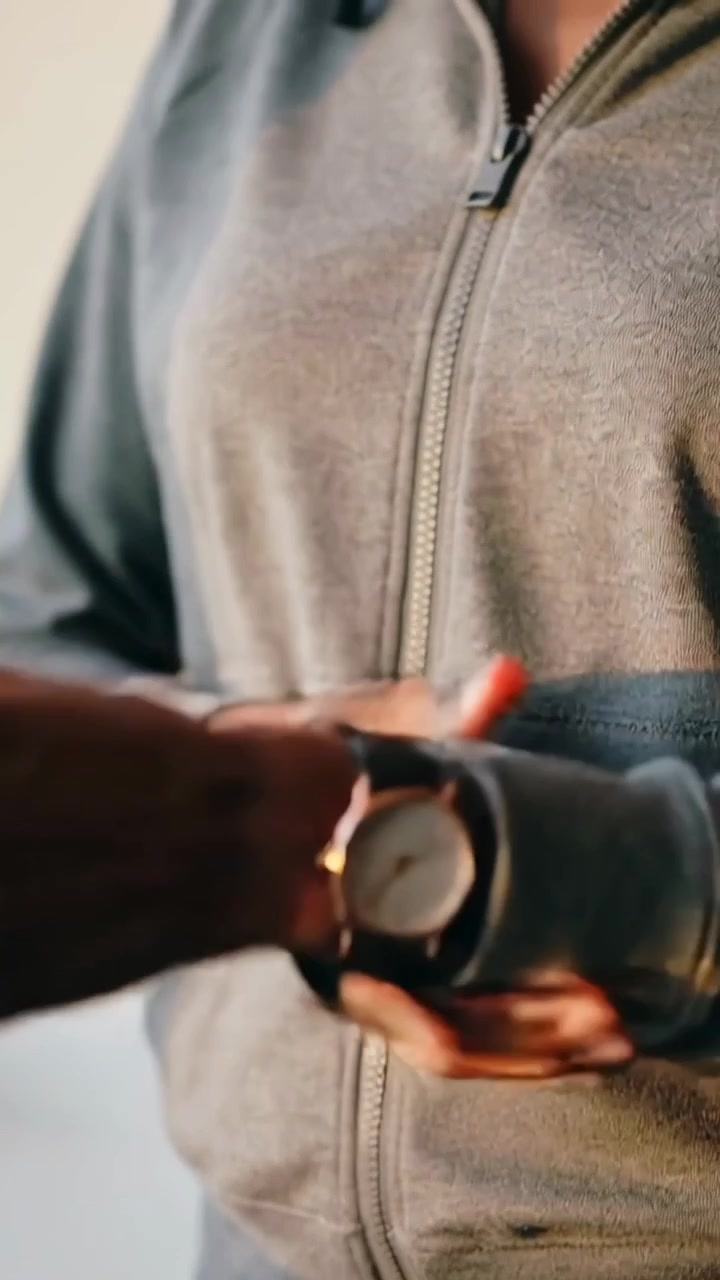 Video verticale: Donna in felpa grigia che allaccia un orologio rotondo sul davanzale di casa, controllandone la vestibilità.