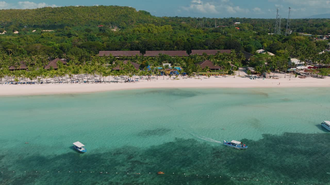 Ampia spiaggia tropicale con mare turchese e sabbia orlata di palme lungo la costa di un resort, camere con vista mare, ripresa aerea in allontanamento, Bohol Filippine.