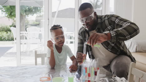 african american father and son sitting at table holding test tubes with liquid, in slow motion
