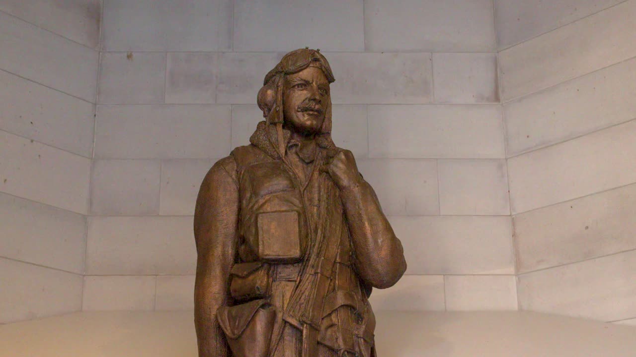 Una lenta inclinazione verso l'alto rivela una statua in bronzo di un pilota militare contro uno sfondo di muro in pietra.