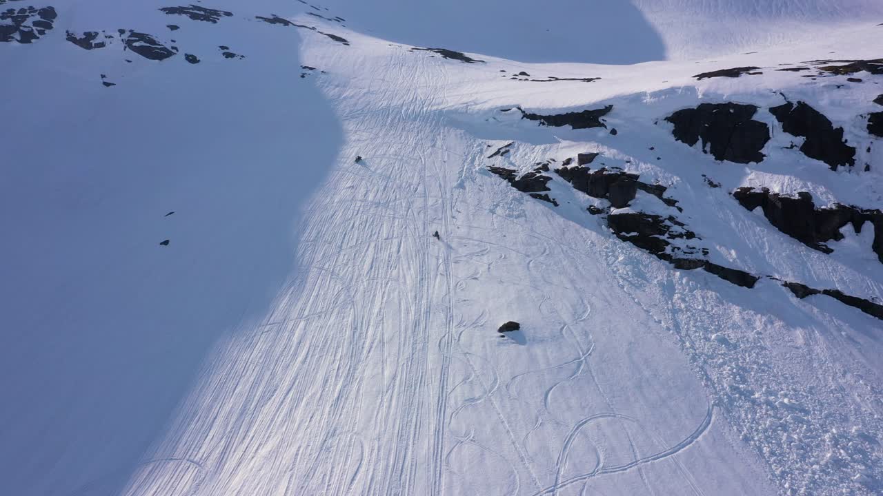 Aerial view over snowy mountain landscape. Following two snowmobiles driving up hill and turning down. Sunny weather. MOVING FORWARD.
