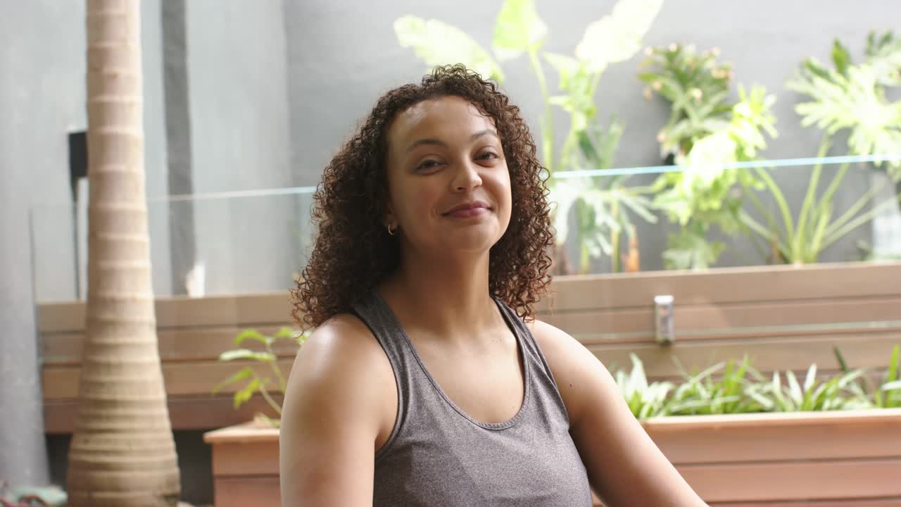 Mujer afroamericana relajándose en el patio con una camiseta gris, inclinándose hacia atrás, respirando profundamente y sonriendo junto a unas macetas