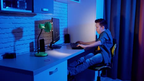 excited boy playing games on a computer at home. teen gamer sitting at the table in his room and playing online video games. blue background.