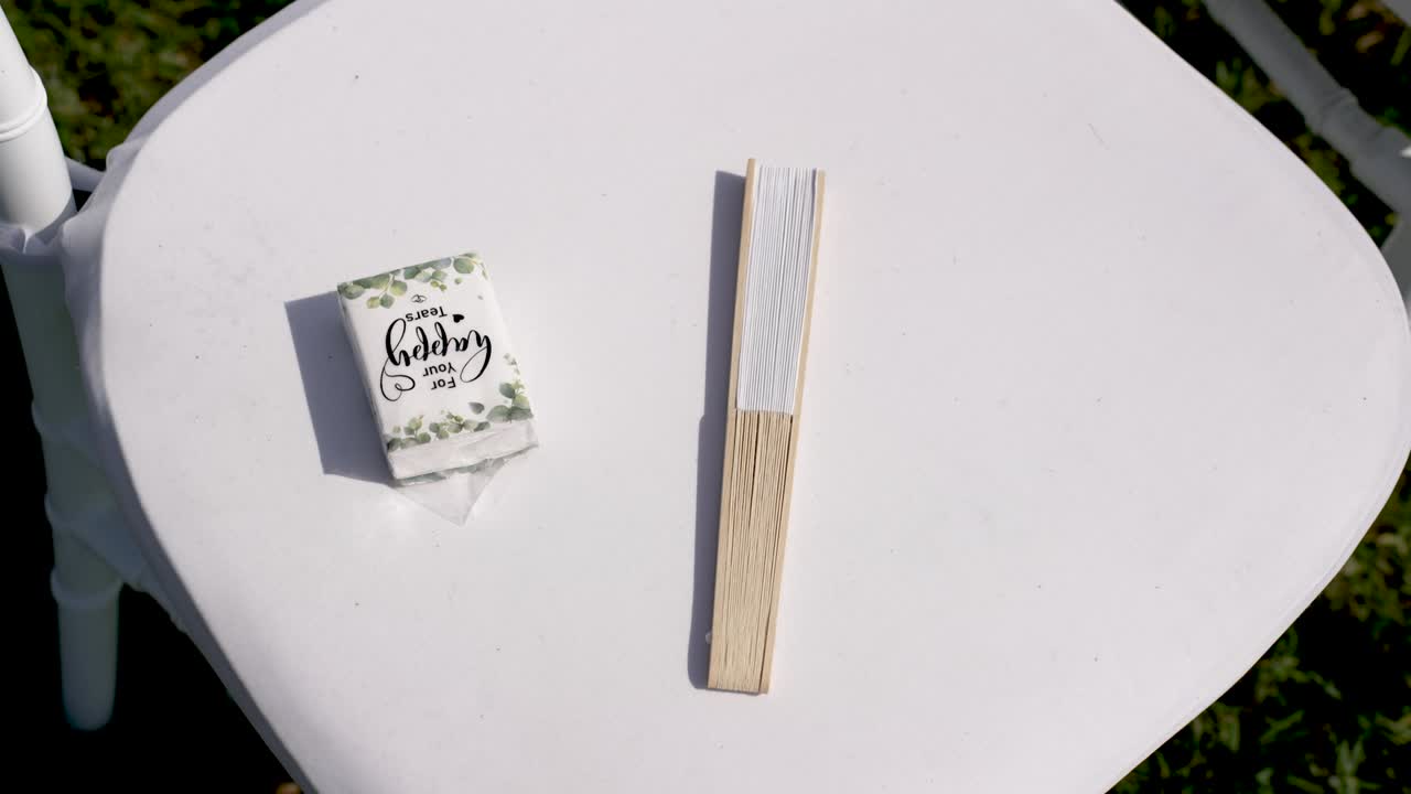 Una piccola scatola di fiammiferi con un design a foglia è posizionata accanto a una ventola di legno su una sedia bianca. L'ambientazione è all'aperto con luce intensa. Gli ospiti sono invitati al matrimonio