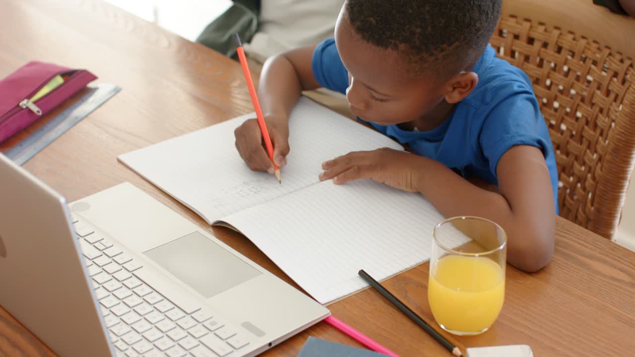 Child is writing homework with red pencil at wooden table, adult entering and placing mug nearby