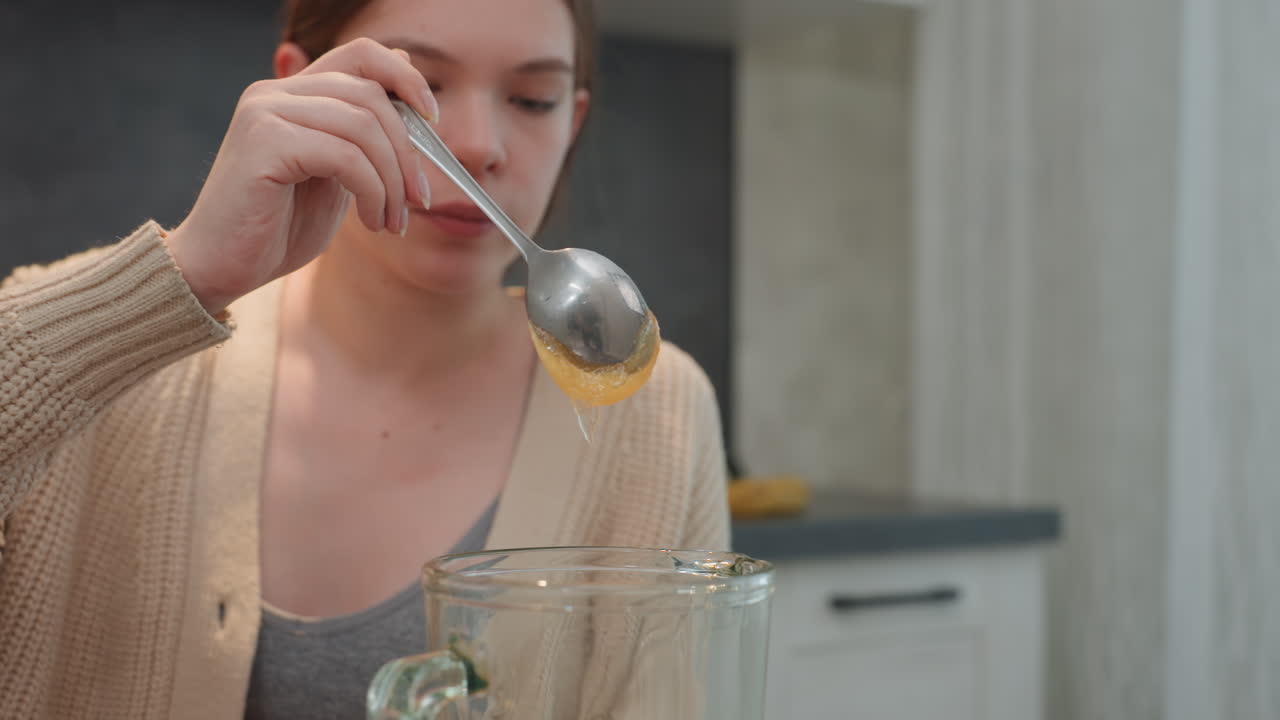 Donna caucasica incinta che misura un filo di miele nel boccale del frullatore in una cucina accogliente, cardigan e bicchieri sul bancone, espressione delicata e concentrata, versamento a cucchiaio, rituale nutrizionale mattutino che enfatizza