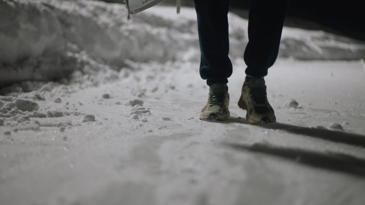 Rear view of person walking on snow-covered surface who steps on ice chunk, causing shoe to collide with icy obstacle, leaving footprints and casting long shadow across cold ground