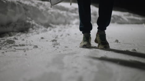 rear view of person walking on snow-covered surface who steps on ice chunk, causing shoe to collide with icy obstacle, leaving footprints and casting long shadow across cold ground