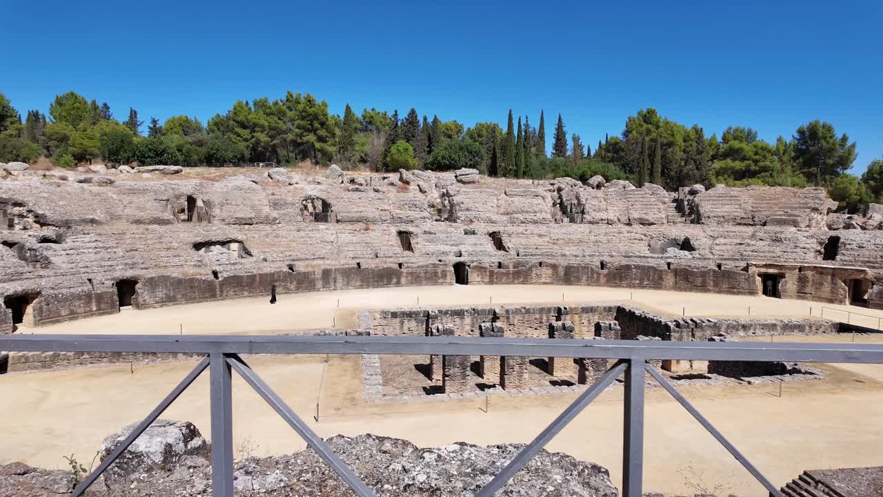 Vista attraverso un arco di pietra grezza che rivela le rovine dell'antico anfiteatro romano nel sito archeologico di Italica vicino a Siviglia, Spagna. Strutture in pietra storiche e architettura classica.