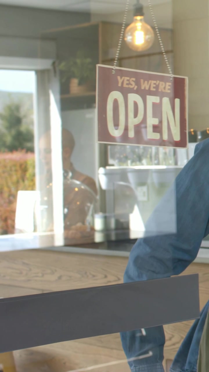 Video verticale: Ingresso in un caffè, uomo con giacca di jeans e grembiule che gira il cartello "APERTO", apertura, spazio per il testo.