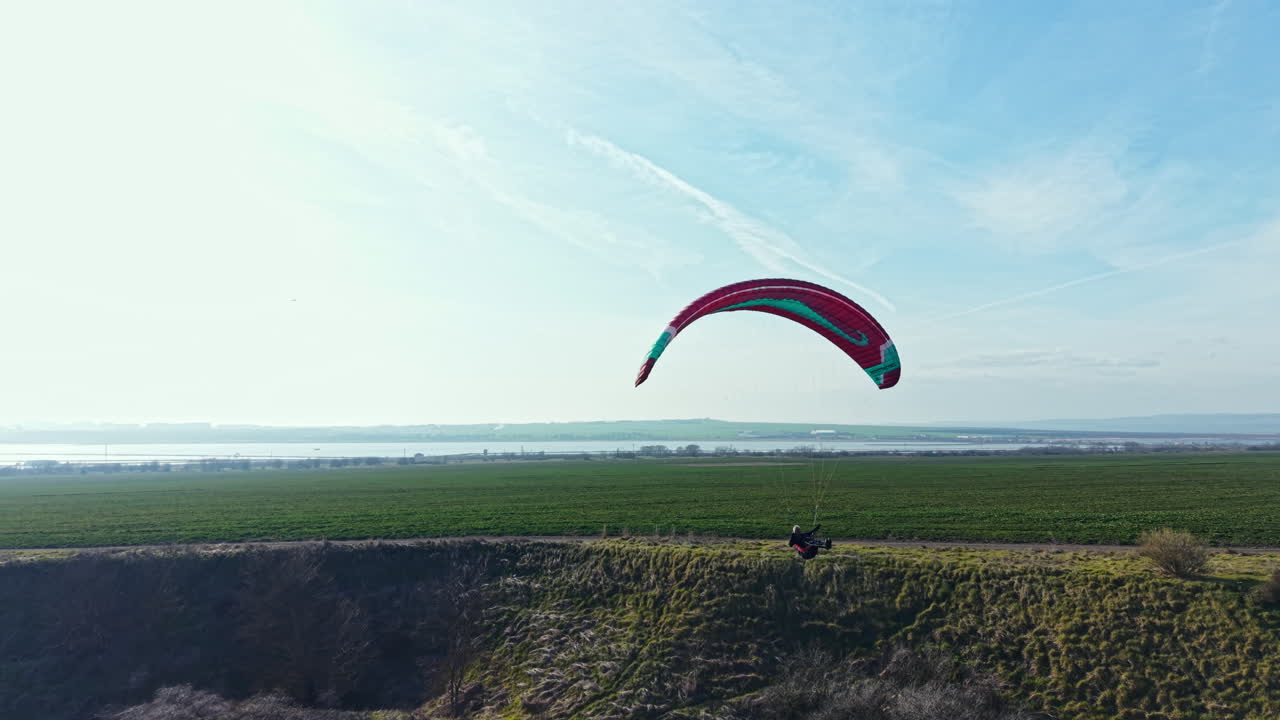 Person flies a paraglider over a green landscape with cliffs and a clear sky during daylight hours in an outdoor setting