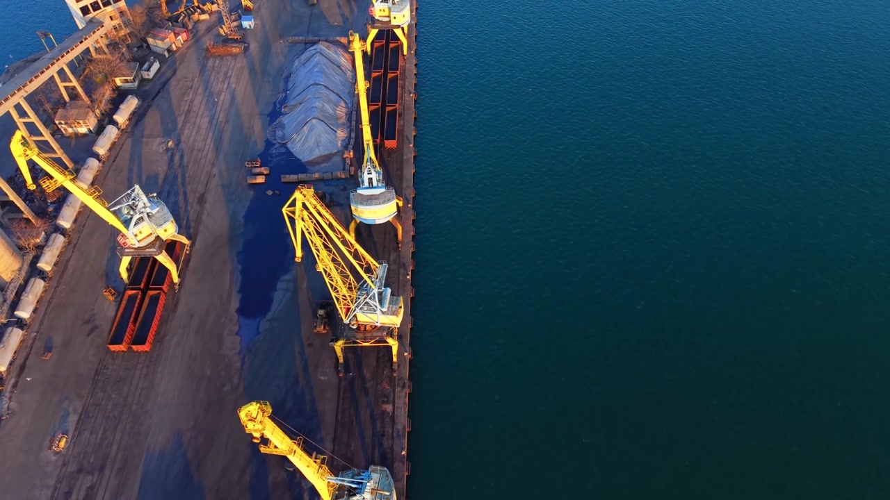 Yellow cargo cranes at a sea dock. Aerial view of heavy industrial machinery standing in a row next to deep blue water with long shadows.