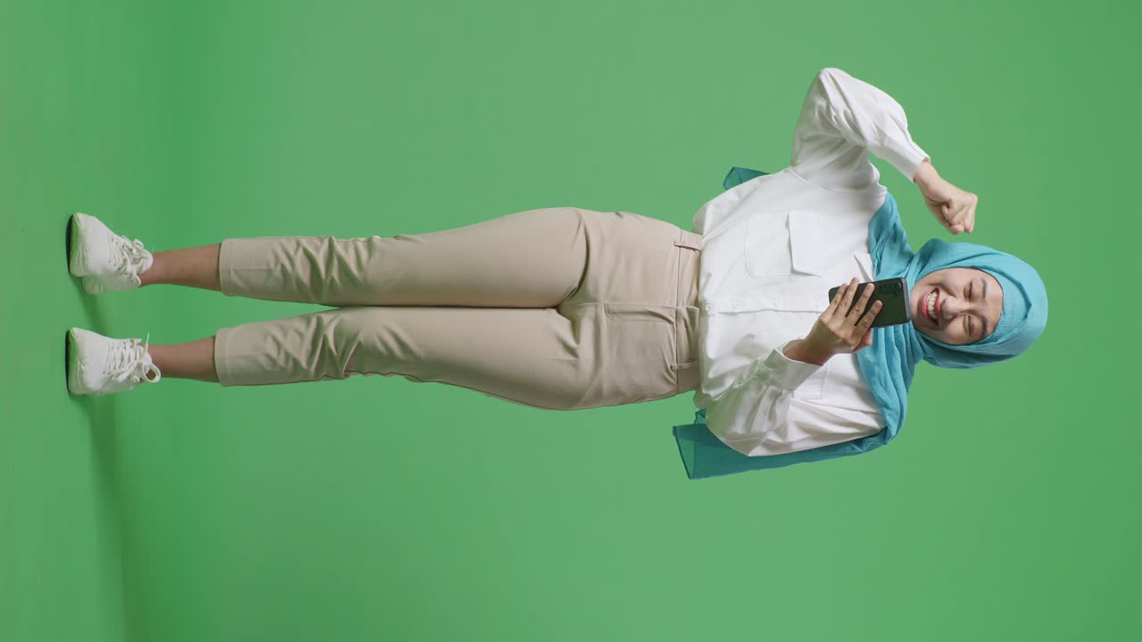 스마트폰을 보는 아시아 무슬림 여성의 몸이 가득 차 있고, 그 다음에는 초록색 스크린 배경 스튜디오에 서서 축하하는 골을 비명을 지르고 있습니다.