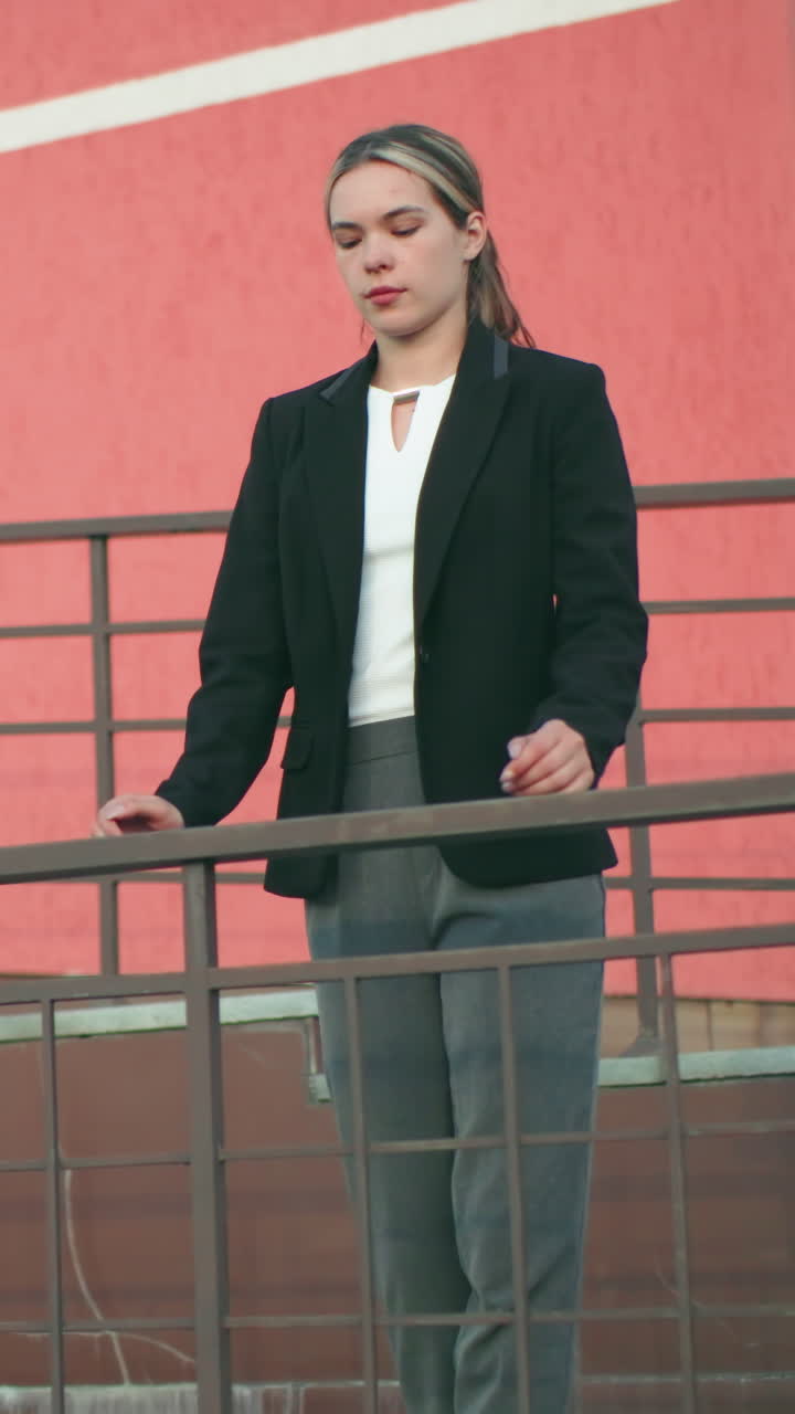 Young champ in black blazer rests hands on iron railing outside residential building painted red, exuding confidence and calm with focus on modern architecture and urban environment in soft lighting