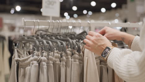 lone shopper hands in ribbed sweater sleeve and gold watch sift beige joggers on metal rack inside brightly lit store, inspecting fabric texture and size tags while deciding casual wear choice
