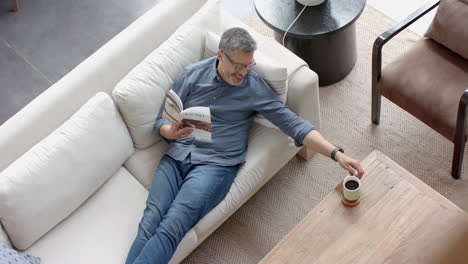 Happy-senior-biracial-man-on-couch-having-coffee-and-reading-book-in-living-room,-slow-motion