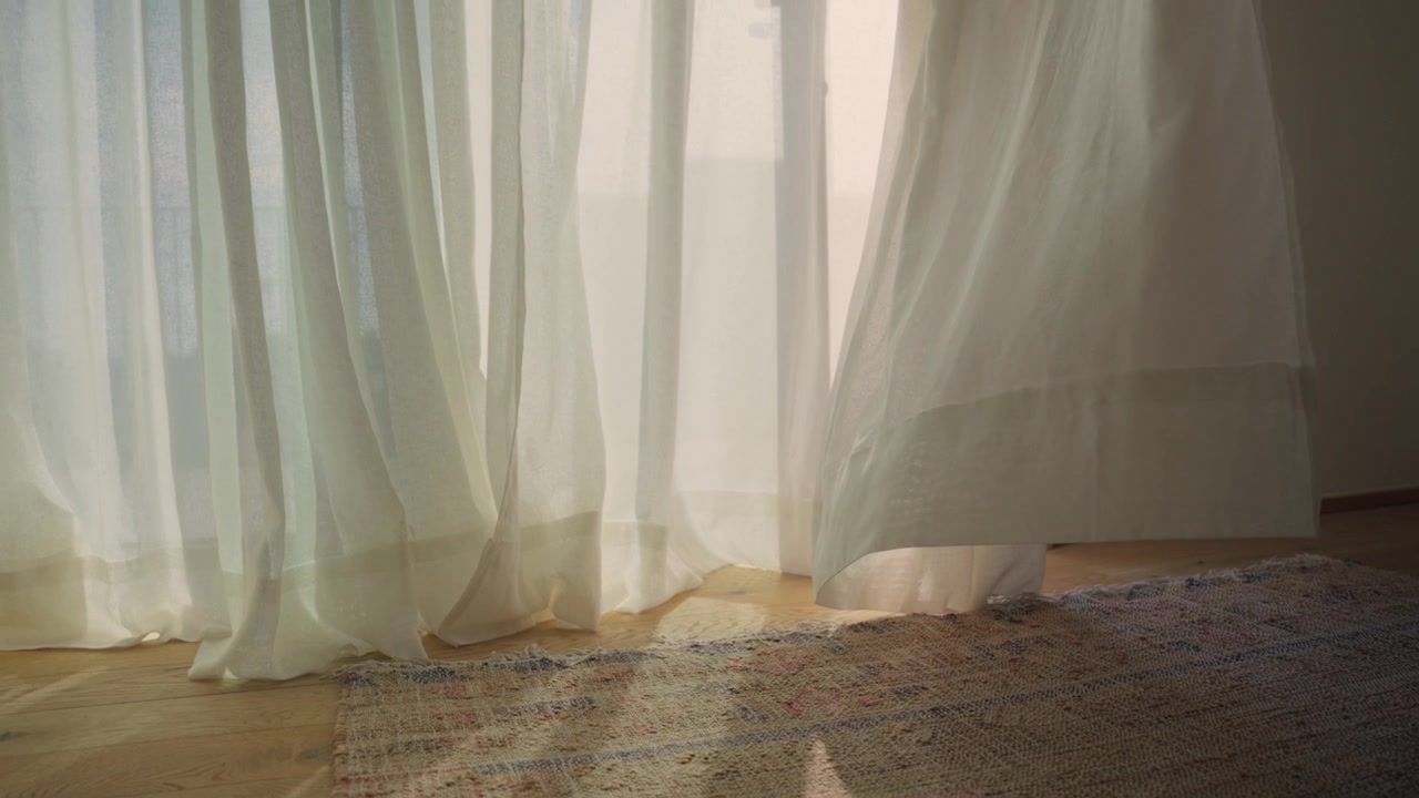 White sheer linen curtains fluttering in the wind against a wooden floor with soft sunlight