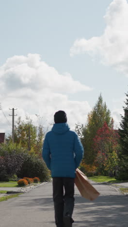 niño con capucha azul y gorra negra camina por la calle residencial con una bolsa de papel, rodeado de follaje de otoño, y casas coloridas bajo cielos nublados