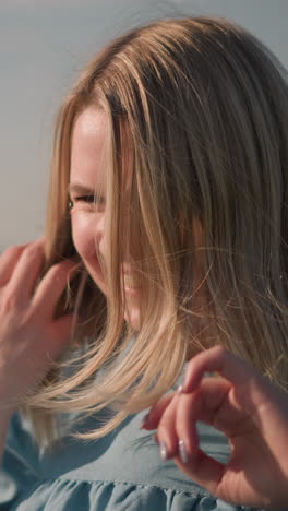 a close-up shot of a woman in a blue shirt as the wind blows her hair. she gently moves her hair away from her face, creating a natural and serene moment with a blurred background