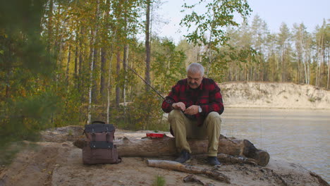 pescador de pelo gris está poniendo cebo en el gancho de la vara preparándose para girar la pesca en la orilla del río en el bosque acampar y hacer senderismo