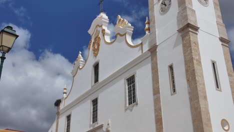 White facade of Portimao Parish Church with golden details and a stork nest on the roof, under a blue sky with some clouds