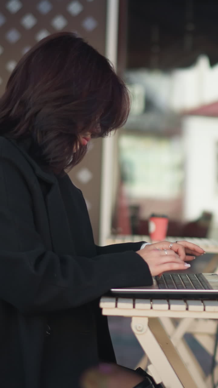 노트북 화면에 초점을 맞춘 여성의 측면 뷰, 자연 빛 아래 도시 환경에서 야외에 앉아있는 동안, 장면은 현대 가구, 건물의 흐릿한 배경을 특징으로합니다.