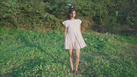 tiro estático en cámara lenta de una joven india bonita vestida con un vestido rosa de pie en el jardín en el césped entre árboles en el fondo mirando a la cámara