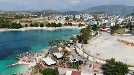 Drone-view-in-Albania-vertical-lift-showing-vacation-town-next-to-the-blue-clear-water-sea-and-white-sand-beaches-in-Ksamil