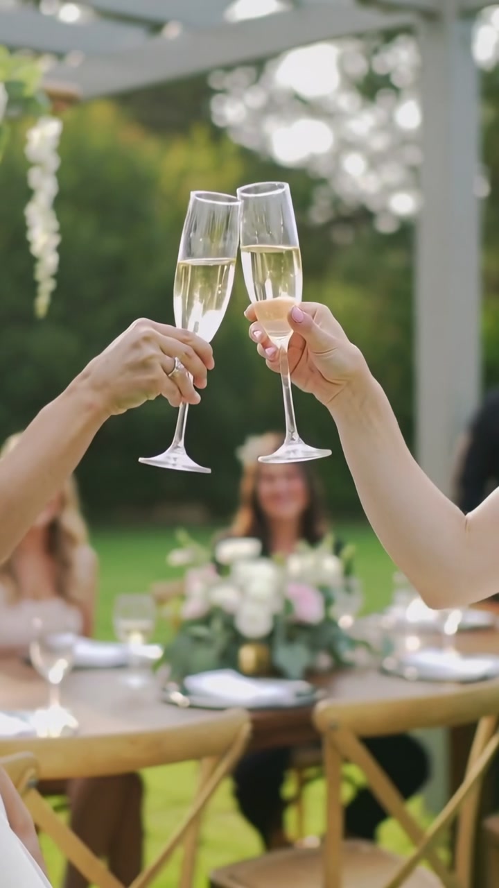 Friends and family in smart attire raising champagne flutes clinking over table on lawn under arbor