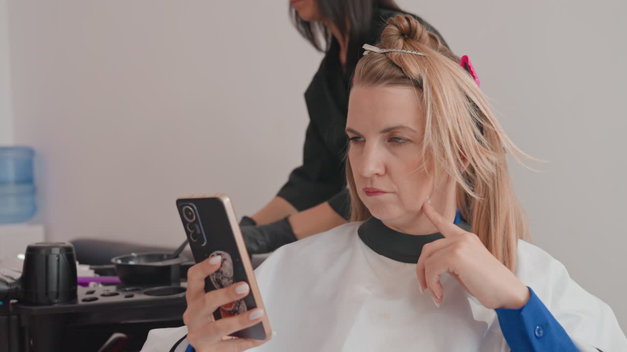 Mujer caucásica en un salón revisando el teléfono; la clienta se sienta con una capa mientras el estilista aplica decolorante y papel de aluminio al cabello trenzado; manos enguantadas ajustan secciones; se usa un smartphone para mensajes y como referencia en el espejo.