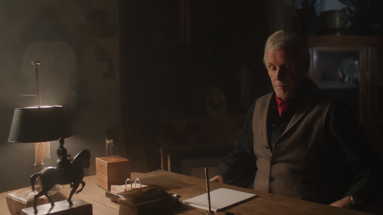 Old passionate man sits down to his desk, put on the glasses and writes a letter