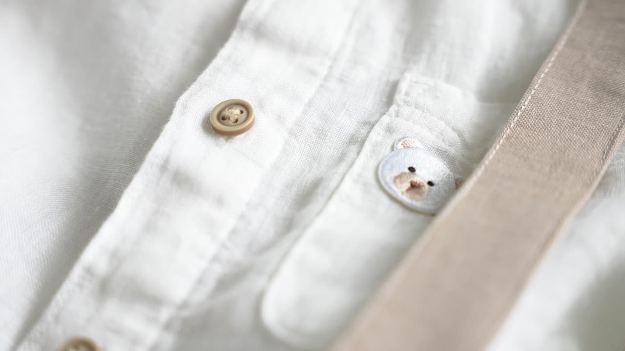 Close-up of a white linen baby shirt with a small embroidered bear face on the pocket, beige suspenders, and detailed wooden buttons, styled for a formal baby outfit or special occasion