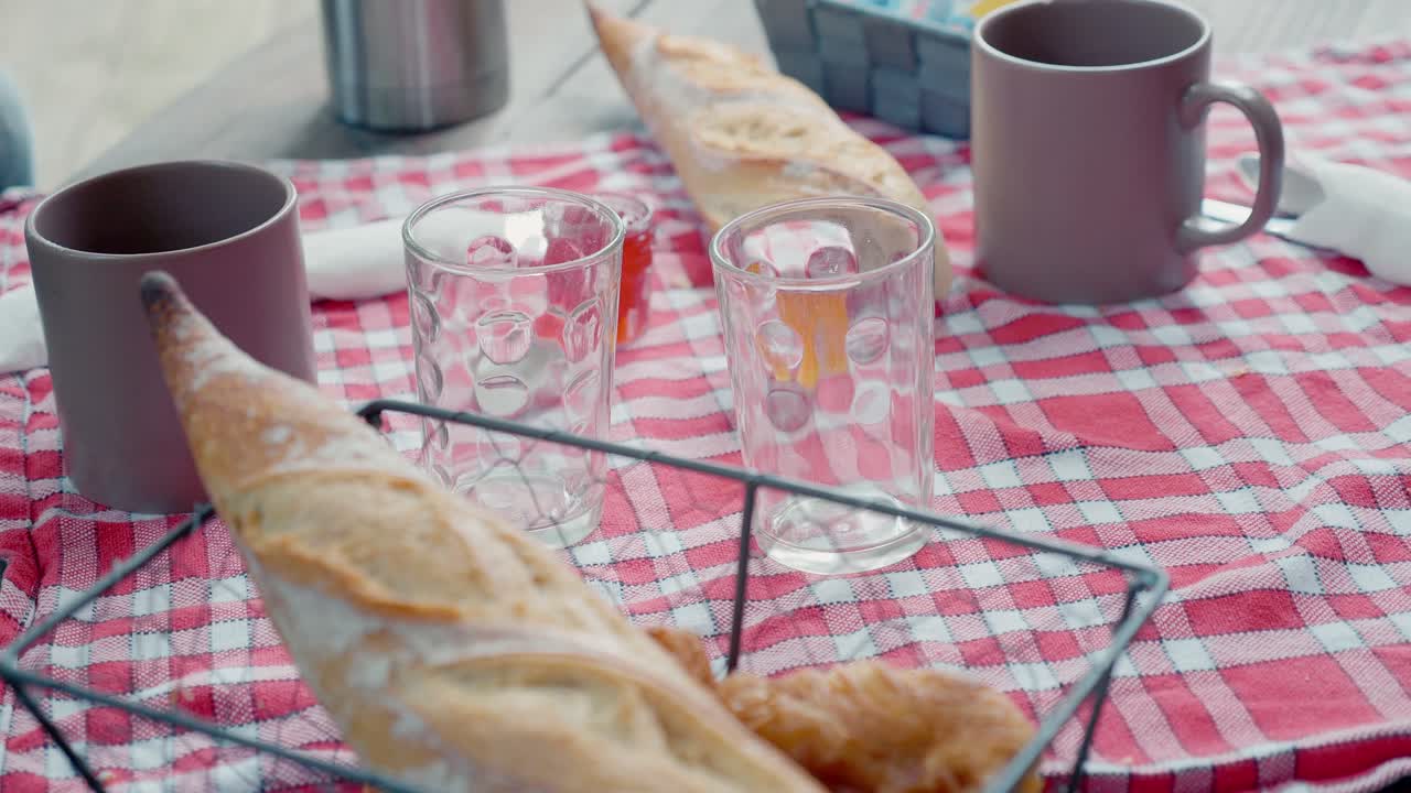 Una baguette fresca posta in un cestino di filo metallico su un tavolo da colazione apparecchiato con bicchieri e tazze vuoti su una tovaglia a quadri rossi all'aperto.