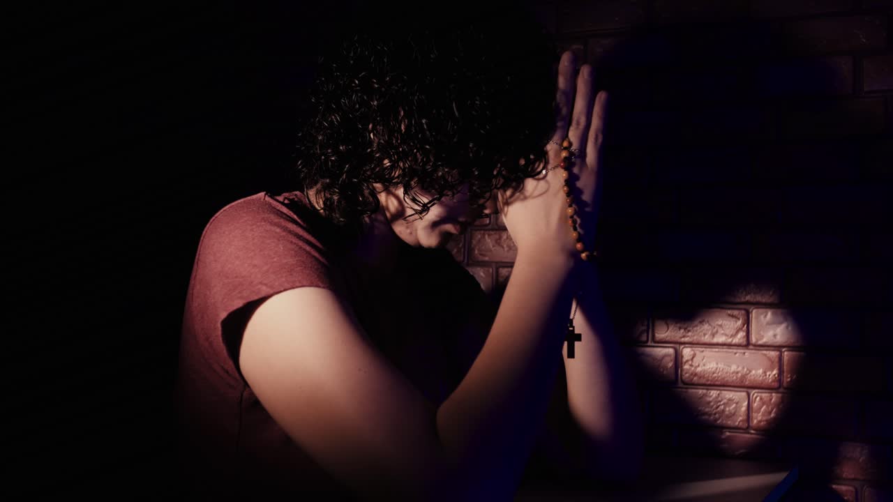 Uomo che prega con il rosario in una stanza buia con luce suggestiva.