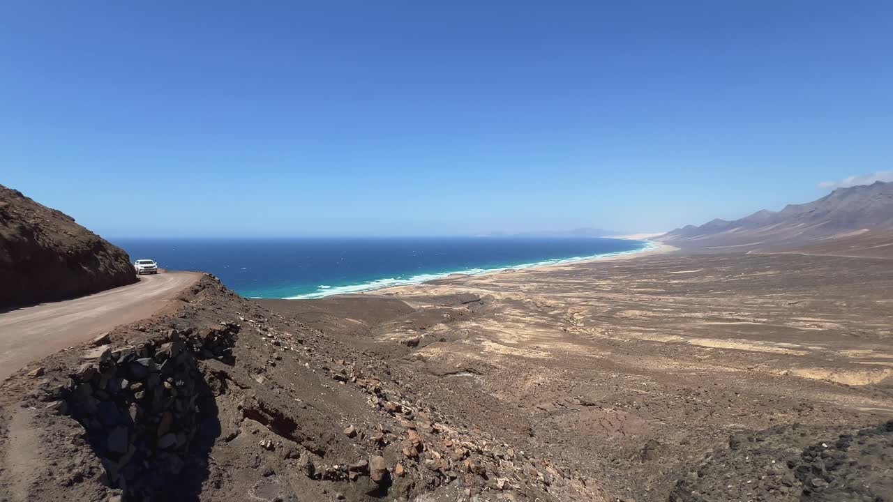 A white SUV car drives on the dangerous, winding dirt road of the Jandia mountain pass, with a breathtaking high-angle view of Cofete Beach. Fuerteventura, Spain