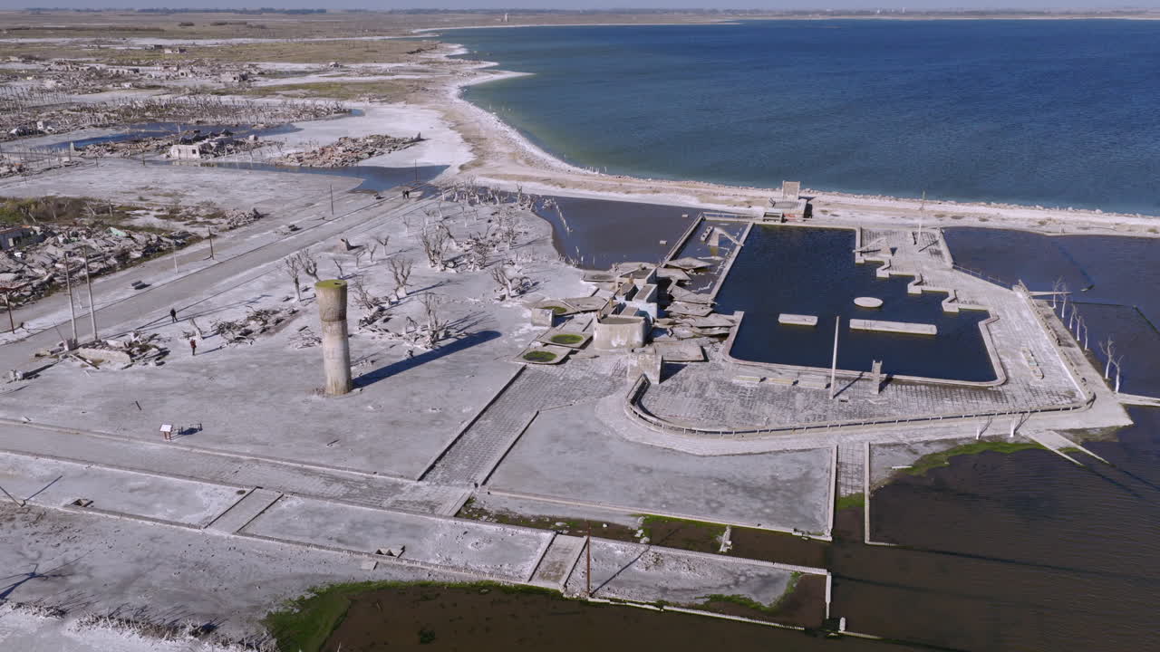 High aerial orbit around the Epecuén ruins, tilted downward to frame the site within the vast lake. Clear day