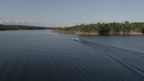 Person-on-a-jet-ski-speeding-along-a-large,-idyllic-lake-along-the-tropical-shoreline---aerial-view