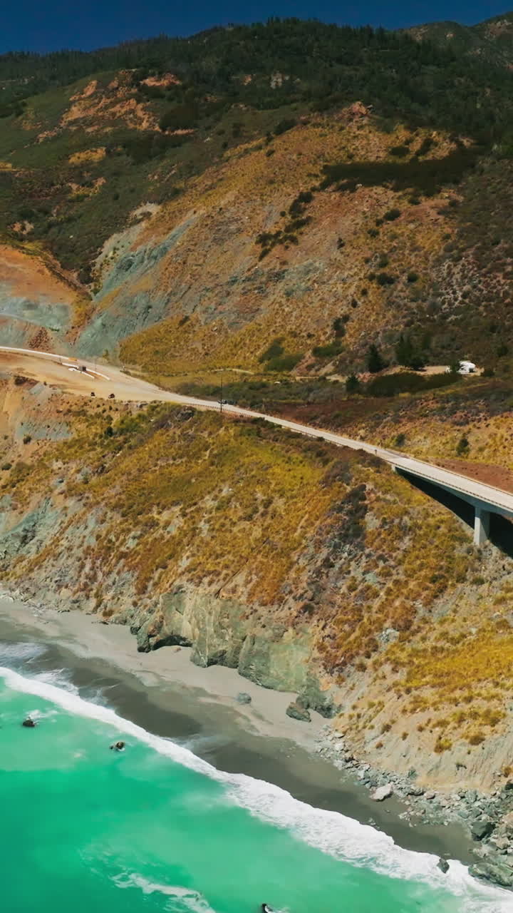 Amazing turquoise water of the Pacific with foamy waves arriving to the rocky shore. Mountains at the coastline with highway and bridge on scorching summer day. Top view. Vertical video