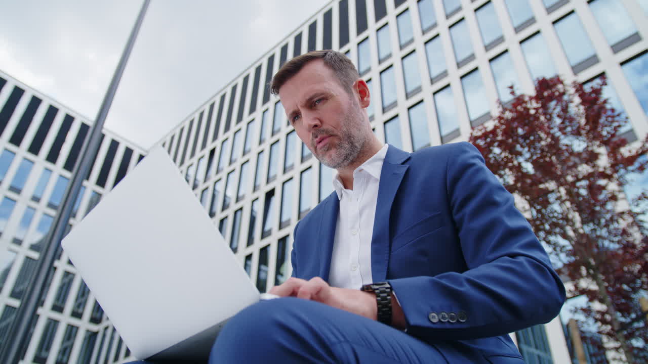 Empresário de terno azul usando laptop ao ar livre, vista próxima do chão.
