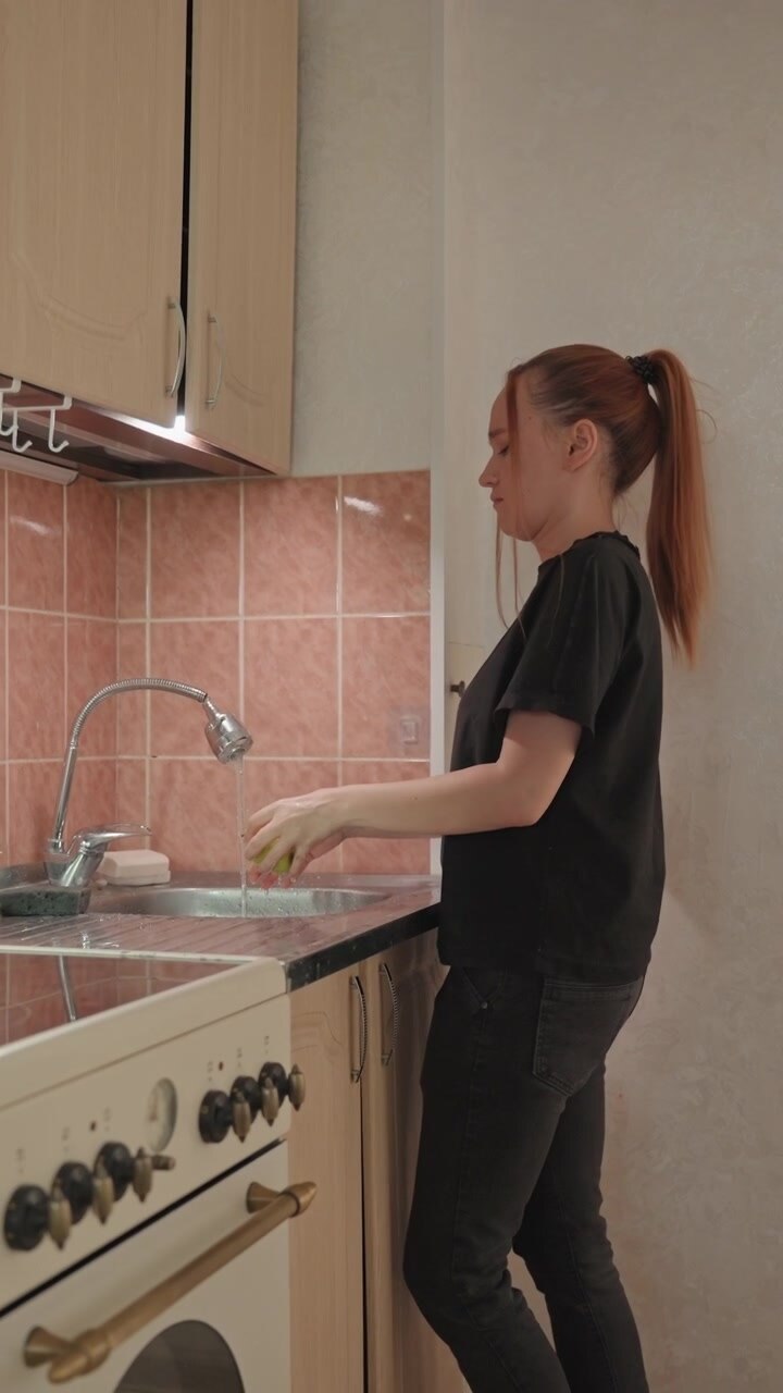 Giovane donna con lunghi capelli rossi legati a coda di cavallo lava un oggetto. La donna indossa una maglietta nera e jeans scuri mentre strofina. Una stufa bianca con maniglie in ottone è visibile a sinistra dell'inquadratura.