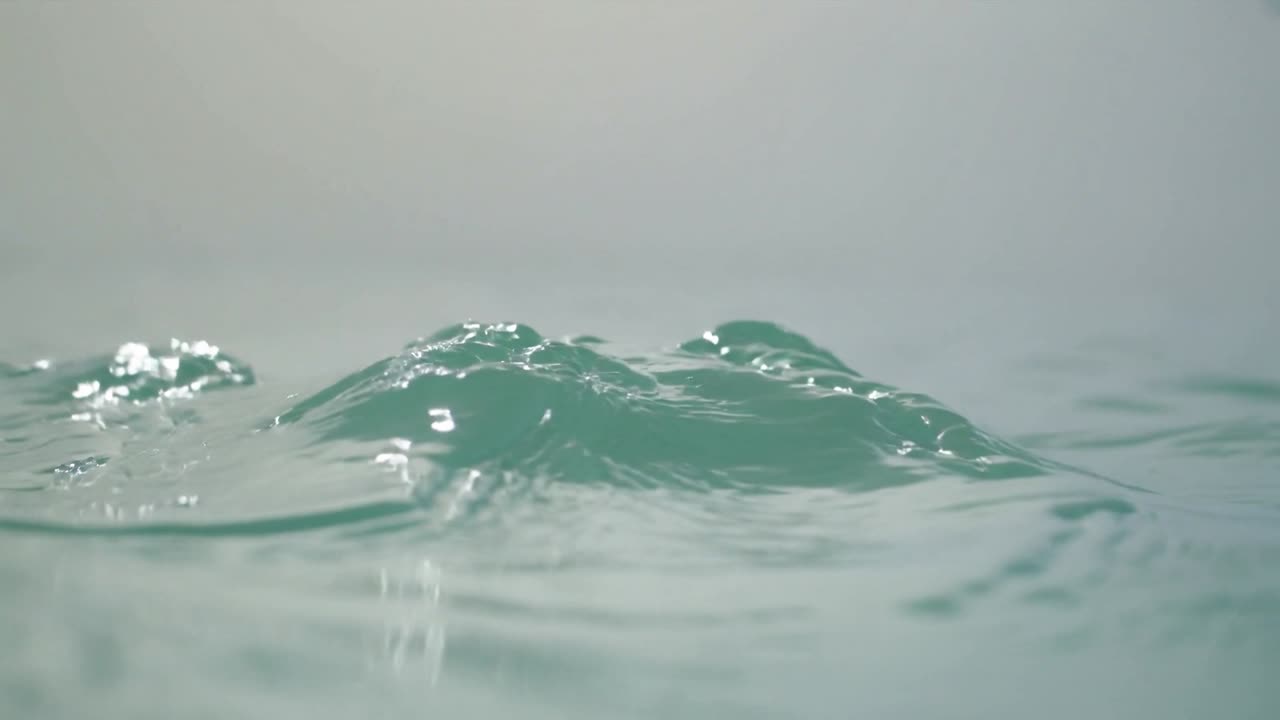 Primo piano della superficie del mare color verde-acqua, con un'onda a sinistra che crea creste lucide e riflessi.