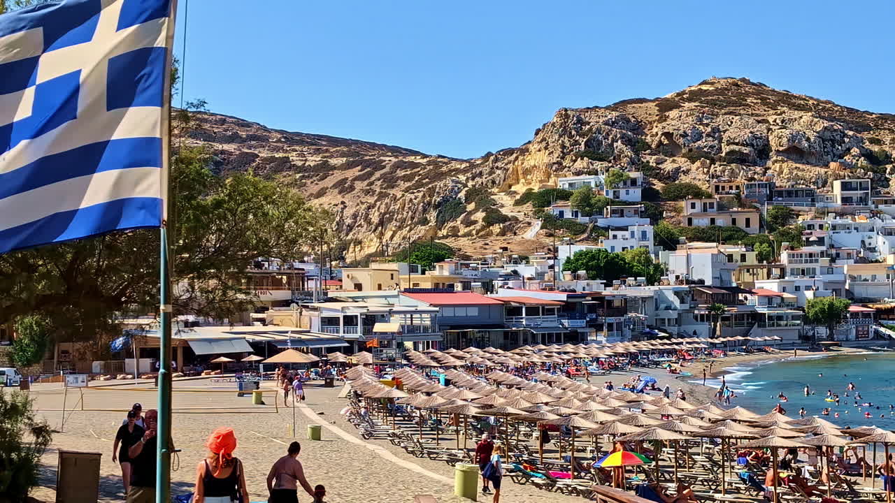 Greek Flag Waving With The Wind With Tourists In The Background At Rethymnon Beach In Greece. - wide shot