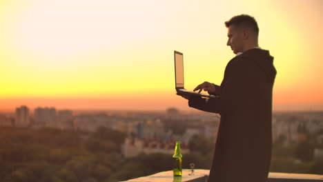 de pie al atardecer en el techo con una computadora portátil y una cerveza. un hombre con capucha trabaja divirtiéndose y contemplando la belleza de la vista de la ciudad desde una altura.