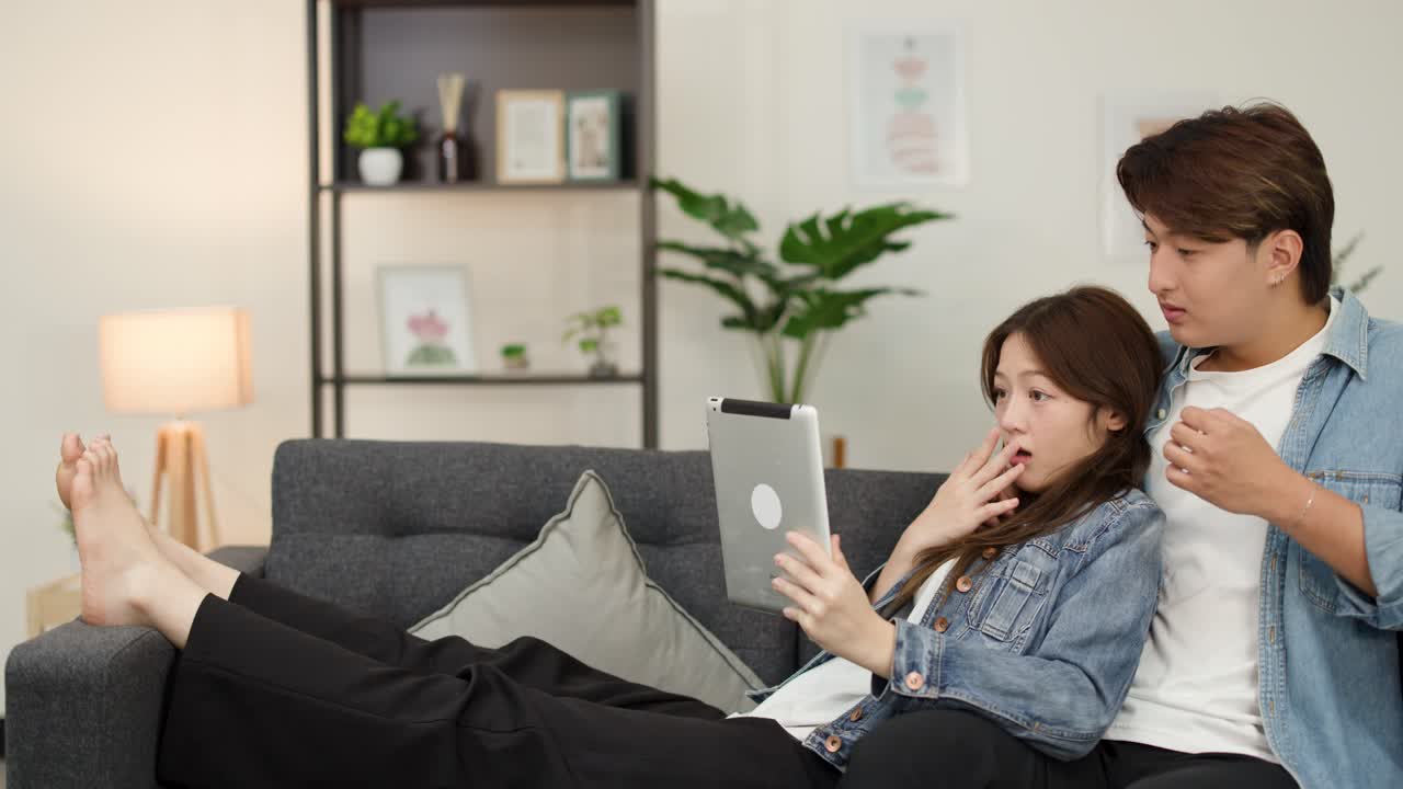 居心地の良いリビングルームで、ソファに座り、恐ろしいタブレット動画を見ている若いアジア人カップル