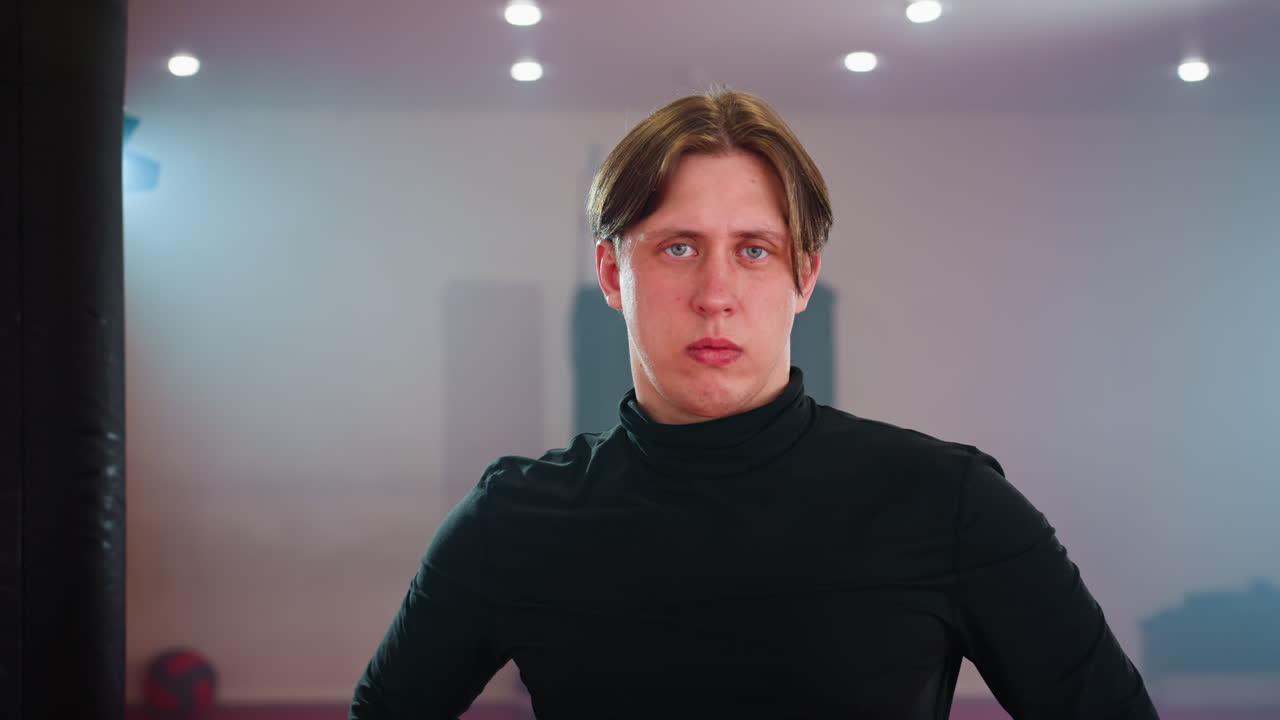 Man in black long sleeve shirt standing in gym with punching bags, hands on hips, appearing focused, serious, calm, and thoughtful during martial arts training
