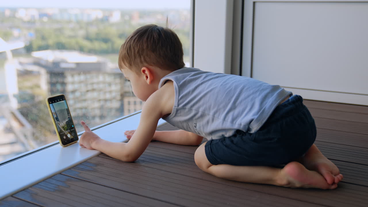 Caucasian toddler sits on his knees touching the smartphone leaning on the window. Little kid taking photos of the street on the phone