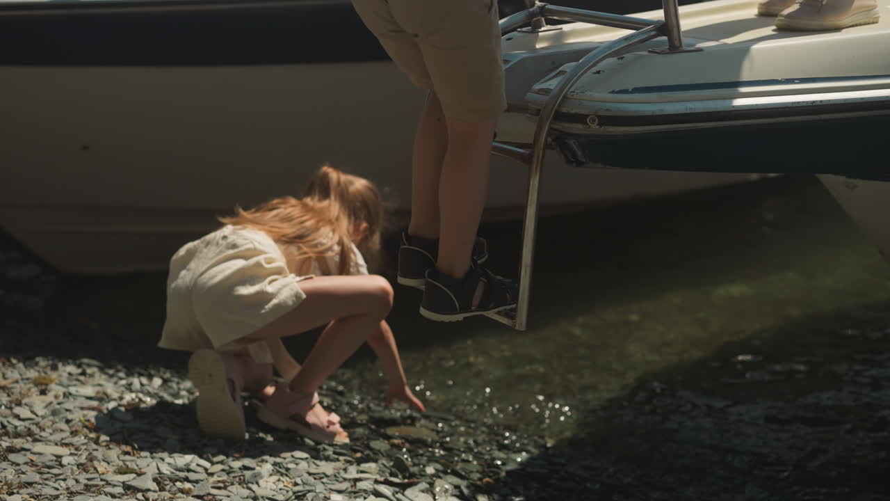 소년은 어머니와 함께 도시 항구에서 금속 사다리를 내려가는 동안 작은 소녀는 자갈로 놀고 있습니다. 가족은 여름 휴가에 관광객 모터 보트를 떠납니다.