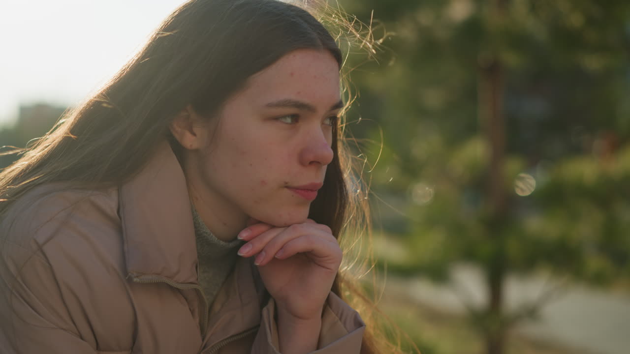 una joven con una chaqueta de melocotón está profundamente pensativa, descansando su barbilla en su mano mientras está sentada en un parque. la luz solar natural destaca su expresión pensativa contra un fondo borroso de árboles
